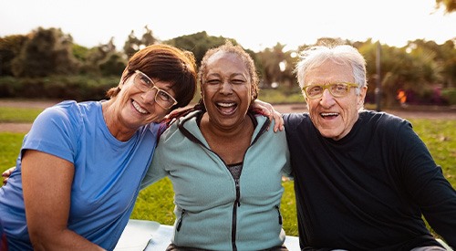 Three adults sitting outside smiling with arms over each others shoulders