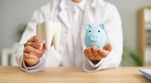 Man in white coat holding large model tooth in one hand and blue piggy bank in the other