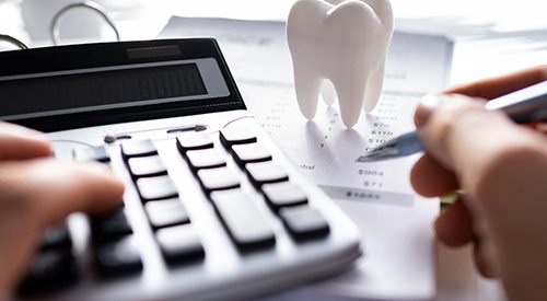 Fingers holding pen and pressing calculator buttons at desk with invoice and model tooth